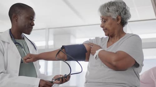 Happy diverse female doctor taking blood pressure of senior female patient in hospital, slow motion