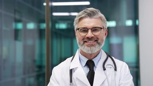 Portrait of smiling mature doctor in white coat and glasses standing in modern hospital clinic look