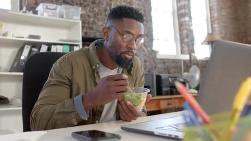 Focused african american casual businessman using laptop and having lunch in office in slow motion
