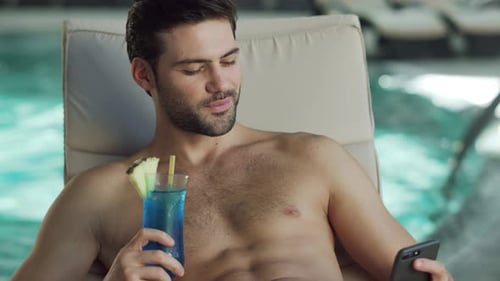 Sexy man enjoys vacation reading news on smartphone by luxury hotel pool
