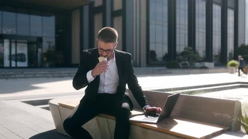 Businessman in Suit Eating Sandwich and Typing on Laptop in Downtown District