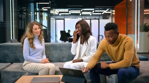 Femmes caucasiennes et noires discutant affaires pendant qu'un homme noir utilise une tablette, assis sur un canapé