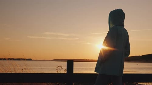 Frau in einer Herbstjacke mit Kapuze bewundert den Sonnenuntergang über dem See