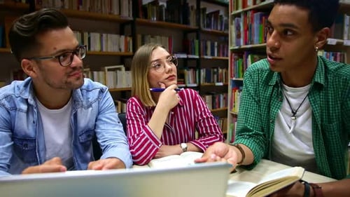 Three young students study in the school library, talking and using laptop for researching online.