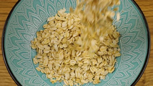 Oatmeal is poured into a white and blue ceramic bowl against a wooden board. Top view. Slow motion.