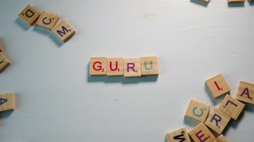 A table with scattered wooden letters and a person spelling Guru. White background. Dramatic light.