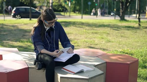 Teenage Girl with Smartphone Doing Homework Sitting in Park Near Street In