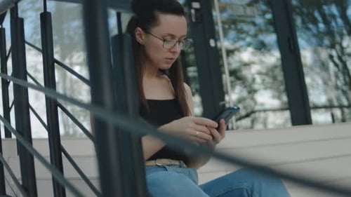 Young Woman with Eyeglasses Sitting Alone on Stairs in Front of a Building Using Mobile Phone