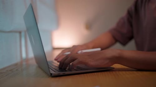 Close-up view of hands typing on a laptop keyboard, in a dimly lit room, conveying a sense