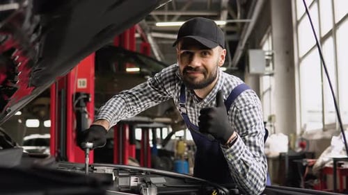 Auto Mechanic Examining Broken Car Engine Standing Near His Car with Open Hood in a Car Repair Shop