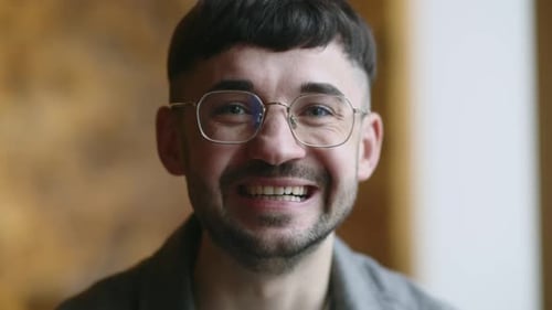Le jeune homme commence à sourire en regardant la caméra : un mec brun riant avec des lunettes sur le devant