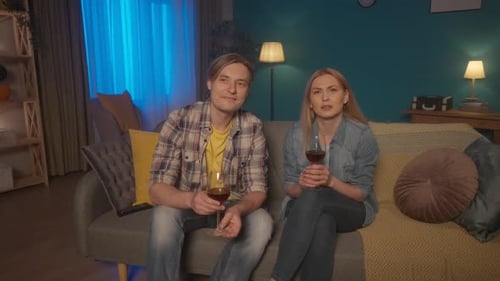 A Young Couple is Sitting on a Sofa with Glasses of Red Wine and Watching TV A Man and a Woman are