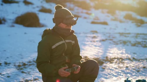 A Person Wearing Goggles and a Beanie Sits on the Snowcovered Ground Holding a Remote Control in