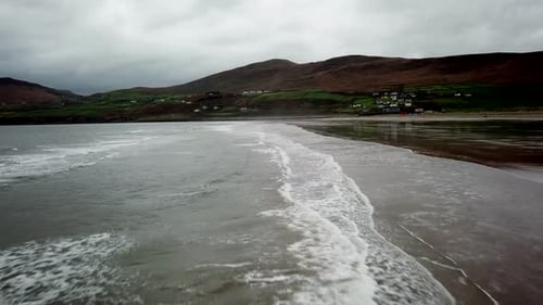 Drone view of a wide Irish beach with mountains behind. Waves wash up on the shore