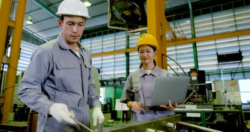 Asian adult male engineer gestures toward machine part while Asian adult female technician checks