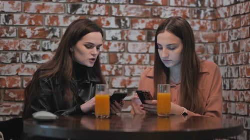 Two Beautiful Girls are Sitting in a Cafe and Talking with Phones