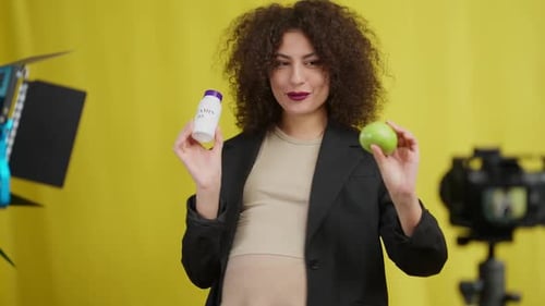 Smiling Pregnant Woman Showing Green Apple and Bottle of Vitamin Supplements Filming Vlog at Yellow