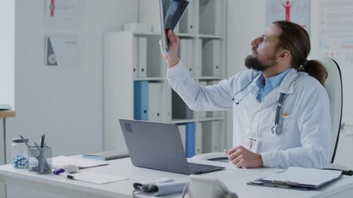 Male Doctor Looking at Chest X-ray and Entering Data into Laptop