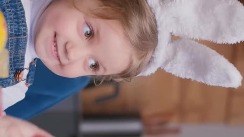 Ein Mädchen mit Hasenohren spielt mit Ostereiern am Küchentisch Vertikales Video
