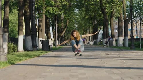 Cheerful Carefree Woman Rides Sitting on a Skateboard Along the Alley with Her Arms Outstretched and