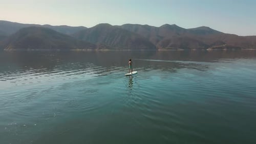 Aerial drone flying over a lake, man doing stand up paddle, with mountains at sunrise. SUP.