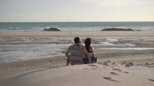 Young couple sit on sandy beach with backs to camera, they embrace and kiss. Slow motion.