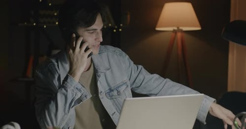 Dedicated Office Worker Young Man Discussing Business on Mobile Phone and Using Laptop at Night