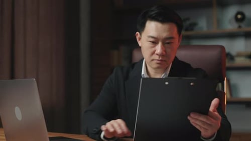 Gentleman Revising Info Between Papers and Laptop in Office