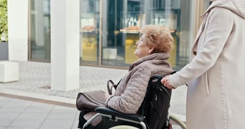 Woman Pushing Senior in Wheelchair Along Urban Street