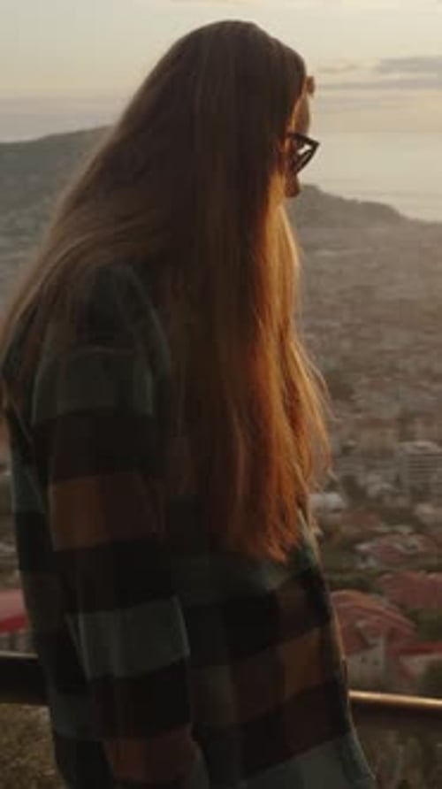 Vertical video. A woman admires the city at sunset on a high mountain, holding onto the railing.