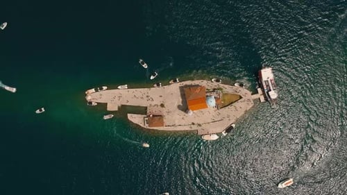 Aerial drone view approaching Our Lady of the Rocks island with a church, tourists, and boats in the