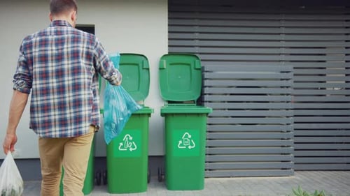 Caucasian Man is Walking Outside His House in Order to Take Out Two Plastic Bags of Trash. One Garb
