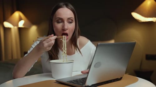 Pretty Woman Eats Instant Noodles and Works on a Laptop