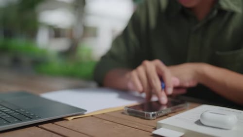 Nahaufnahme eines Geräts, asiatischer Mann schreibt Dokument und schaut auf Handy, Laptop vor sich o