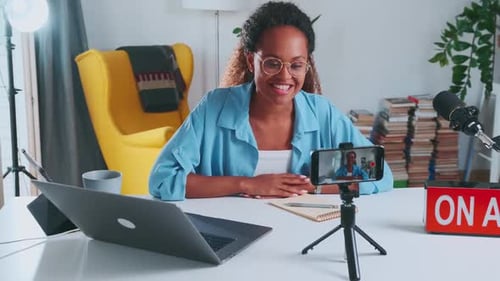 Young African American Woman Blogger Shoots Video on Phone Sits in Home Office