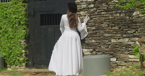 A girl dressed in antique bridal attire, her silhouette framed by the fort’s gates.
