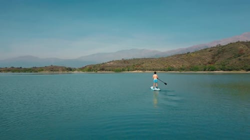 Aerial drone flying over a man doing stand up paddle in a blue water lake with mountains