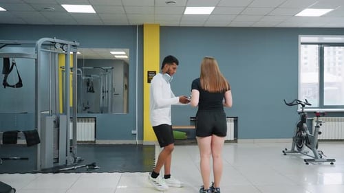 Trainer Hands Dumbbell to Woman Preparing for Knee Lunge Workout in Modern Gym