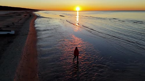 Una mujer camina por una playa de arena, al borde del océano, se puede ver una fuerte puesta de sol en el