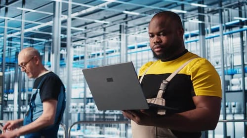 Ingeniero sonriente en planta industrial controla maquinaria automatizada usando una computadora portátil