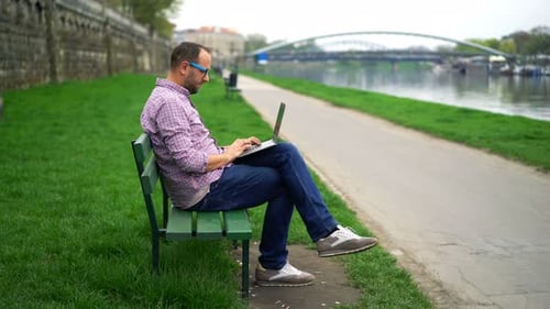 Young man works on laptop at riverside park bench on sunny day