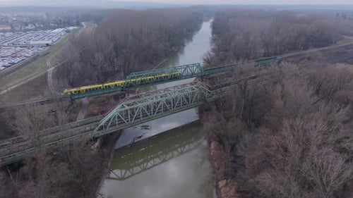 Aerial view of a yellow passenger train crossing a steel railway bridge over a calm river