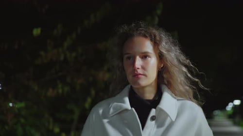 Mulher séria andando ao ar livre à noite vestindo casaco branco sob iluminação suave