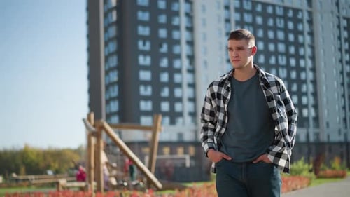 Student with Hands in Pockets Walks Thoughtfully Near Tall Buildings and Playground