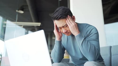 Young exhausted, overworked office worker sitting in the workplace with a severe headache massaging