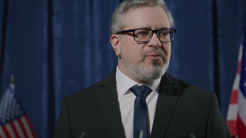 Man in Suit Speaking at Podium with Flags