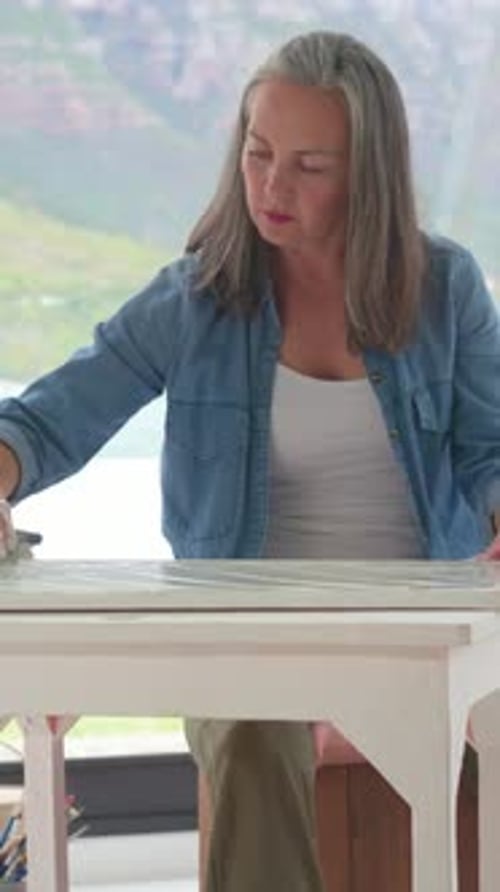 Vidéo verticale : Placement d'une toile, femme étalant un enduit avec un grattoir sur une table blanche, en chemise en jean