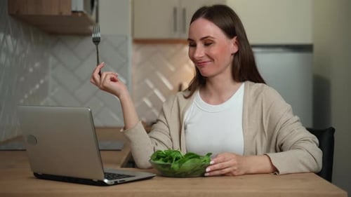 A Woman is Sitting at a Table in the Kitchen and Enjoying a Nutritious Salad for Lunch and Using a
