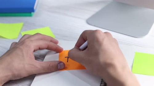 Man writing word Task on orange sticky note at light wooden table, closeup. Planning and deadline