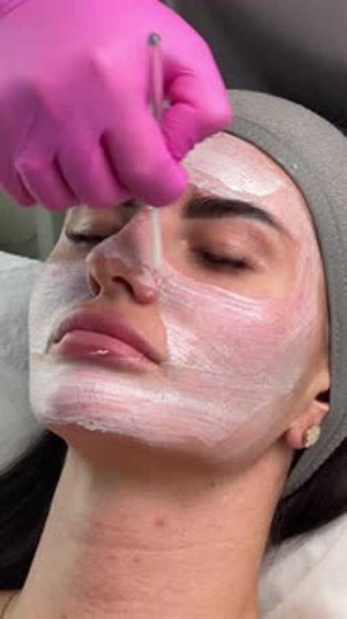 El esteticista aplica una máscara facial blanca en el rostro de una mujer con un cepillo durante el tratamiento de cuidado de la piel